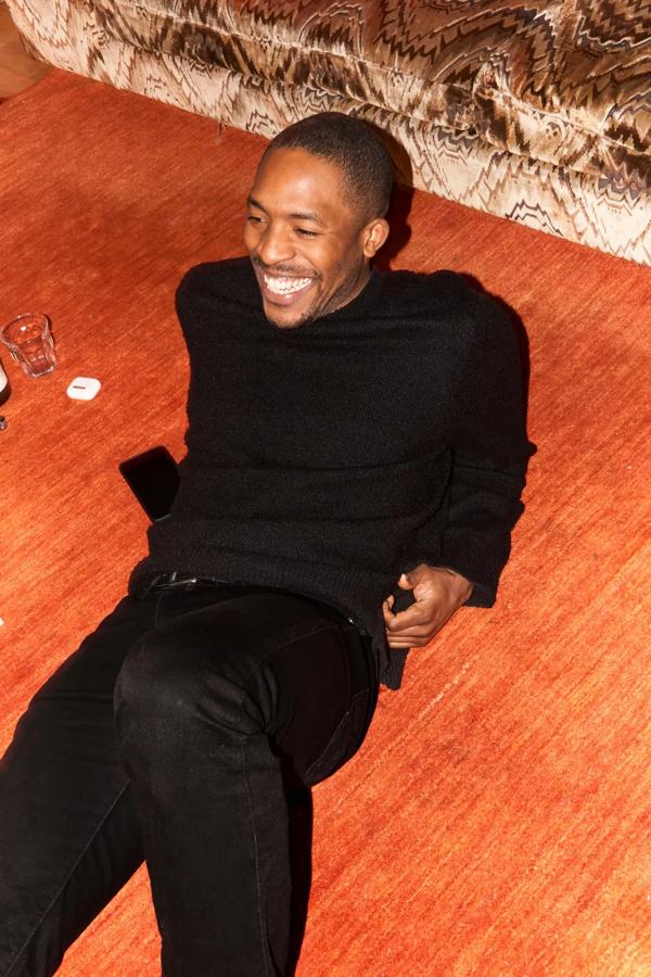 James Massiah wearing black jeans and black jumper sat smiling on wooden floor