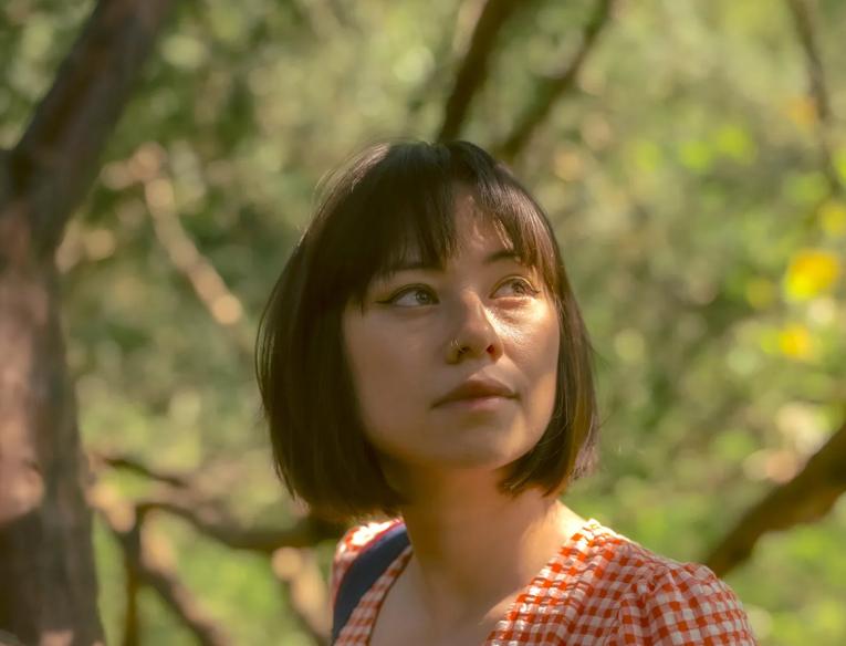 Tori is photographed in dappled light amongst trees. She has short brown hair and wears an orange gingham dress with short sleeves. 