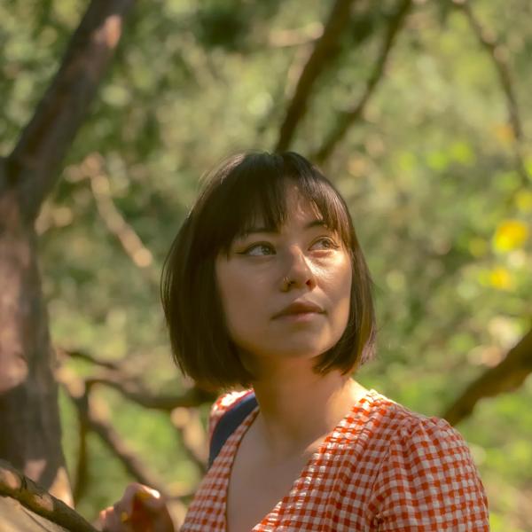 Tori is photographed in dappled light amongst trees. She has short brown hair and wears an orange gingham dress with short sleeves. 