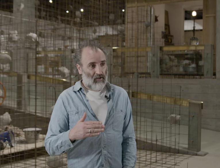 The artist Mike Nelson, a white man with a grey beard, is interviewed whilst standing in the Hayward Gallery, behind him sculptural works from his exhibition Extinction Beckons are visible
