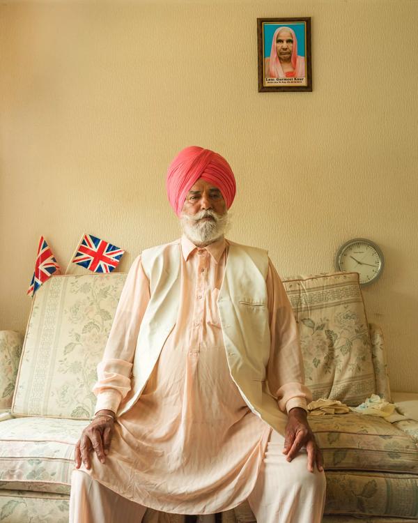 Bhukan Singh is sat on a sofa, wearing a pink turban sat underneath a portrait of his late wife 