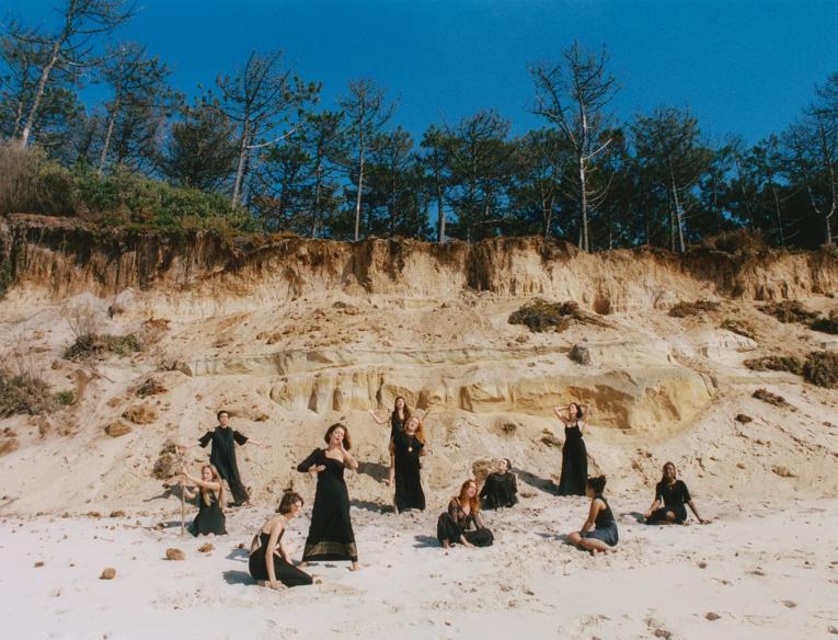 The NYX Choir wearing black performing on a sandy beach with trees behind them.