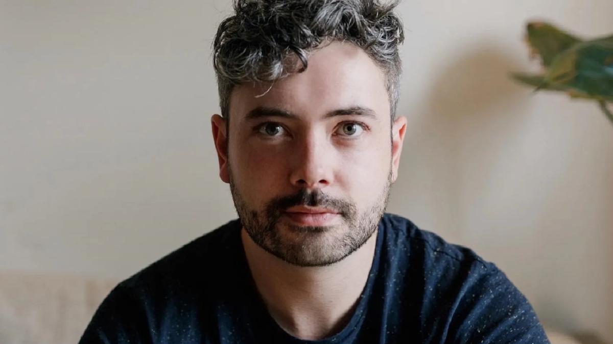 Head & shoulders portrait of poet Seán Hewitt, who has grey tousled hair and wears a dark top with flecks