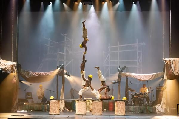 Five performers in construction costumes with yellow hats onstage in a human pyramid