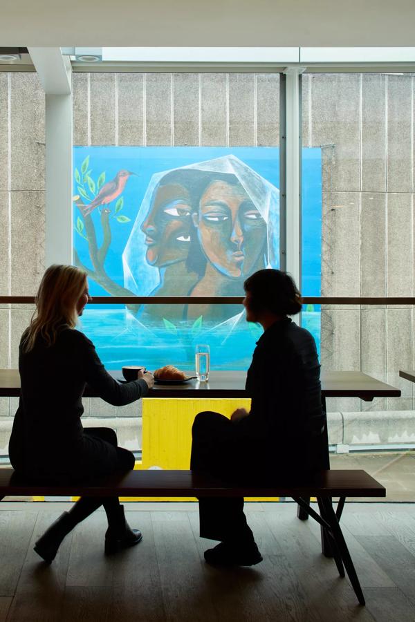 Two customers sit inside the Hayward Gallery Cafe facing a large glass window that looks onto wall art at the Southbank Centre