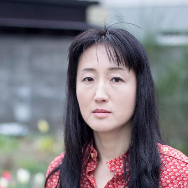 A woman with long black hair staring directly into the camera. She is wearing a red flowery shirt