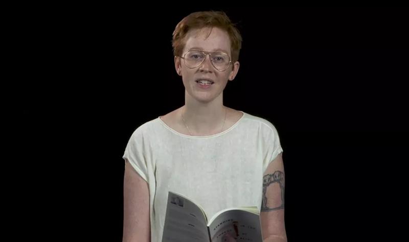 Katie Farris a White woman with short red hair and glasses, wearing a white t-shirt reads from their TS Eliot Prize shortlisted collection against a black background