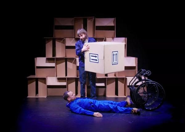 Person wearing a blue boiler suit lying on the floor next to an upturned wheelchair. A person stands above them holding a cardboard box.