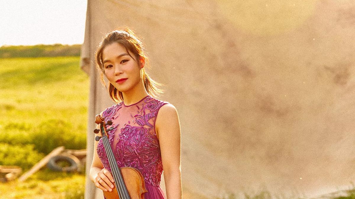 Violinist Esther Yoo in a pink floral dress on a beige backdrop with green landscape in the distance