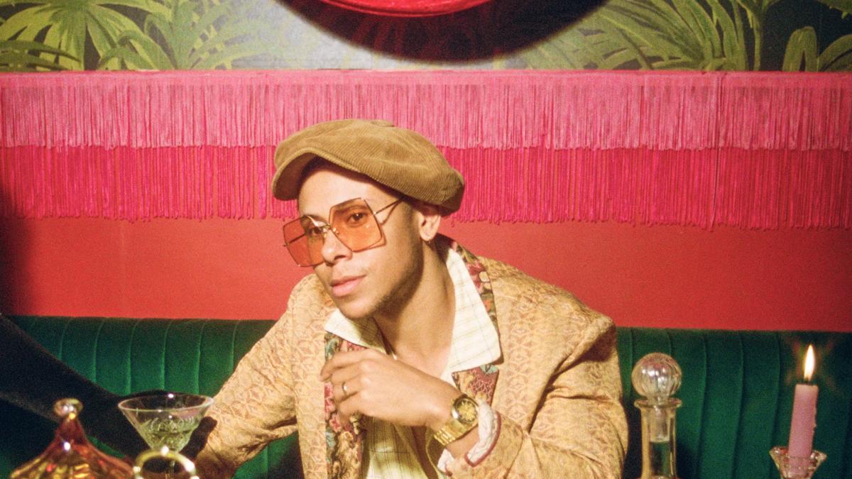 Musician Reuben James sits at a table laden with fruit, wine and crockery. They wear a light beige suit jacket, large square sunglasses and a tan flat cap.