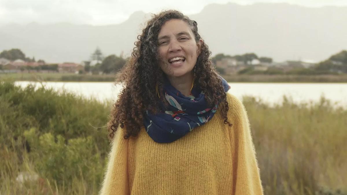 Poet Toni Giselle Stuart stands in wetlands; water and grass is visible behind her, she has long curly hair, wears a yellow sweater and a blue shawl