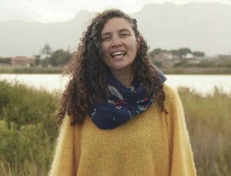 Poet Toni Giselle Stuart stands in wetlands; water and grass is visible behind her, she has long curly hair, wears a yellow sweater and a blue shawl