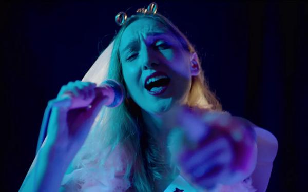 Person singing into a microphone and pointing at the camera, whilst wearing a wedding veil. The lighting is purple and blue and there is a dark background.