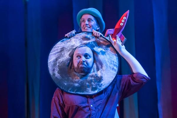 Man with a moon shaped head dress holds a rocket above his head and a woman in a blue bowler hat stands above him.