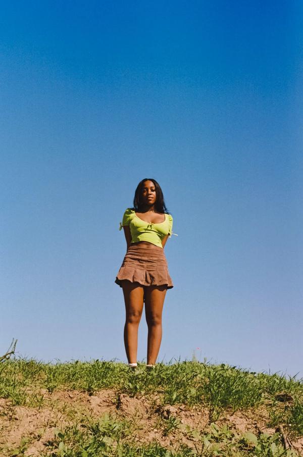 Portrait of artist Rachel Chinouriri on a grassy mound in front of blue sky.