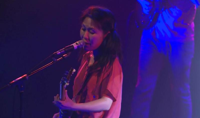 Deerhoof's lead singer on stage at the 2013 Meltdown Festival; she wears a red shirt and plays guitar whilst singing into a microphone