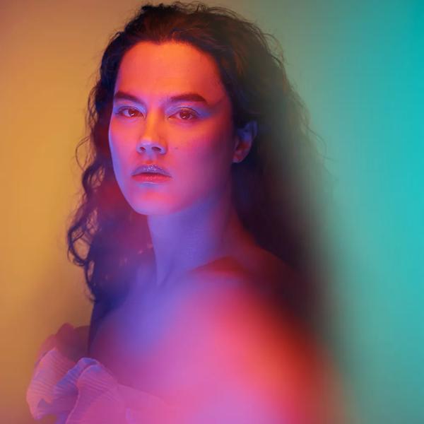 Hazy image of artist Mantawoman wearing a ruffled top and lit with coloured light. 