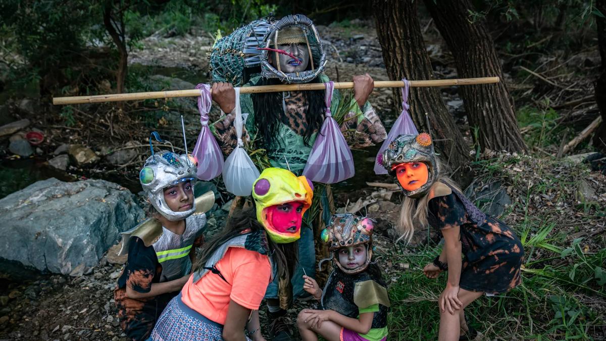 A women in a mask holding a wooden pole with plastic bags hanging off. There are 4 children in helmets crouching in front.
