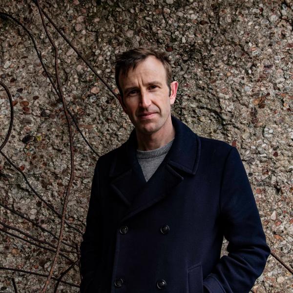 Robert Macfarlane stands against stone background wearing a navy blue coat