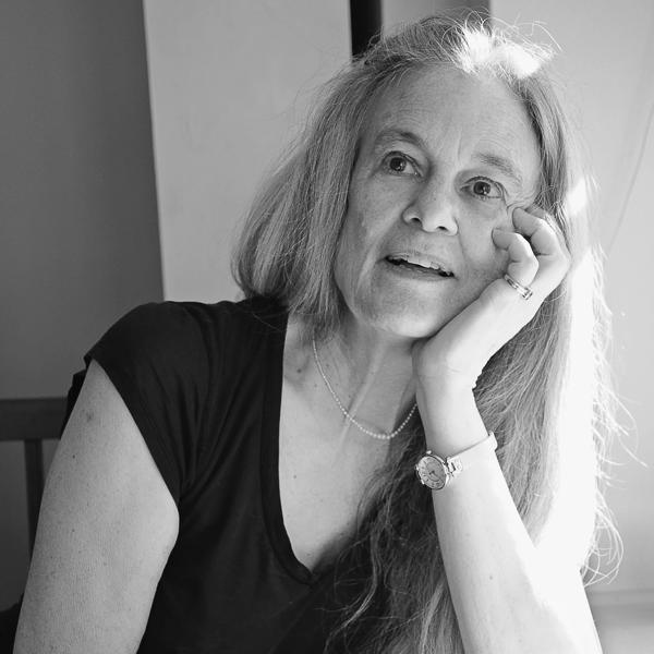 The poet Sharon Olds, an older White Woman with long fair hair wears a dark top and rests her chin on the palm of her left hand