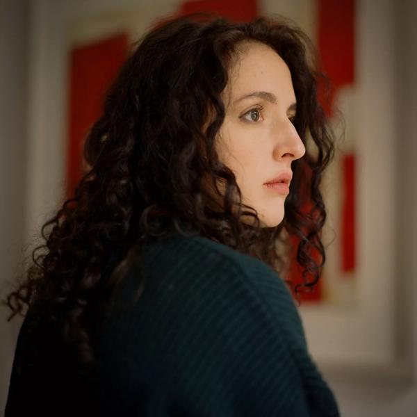 Isabella has dark curly hair and brown eyes. She is wearing a wool coat and pictured in profile against a backdrop of a framed white and red painting. 