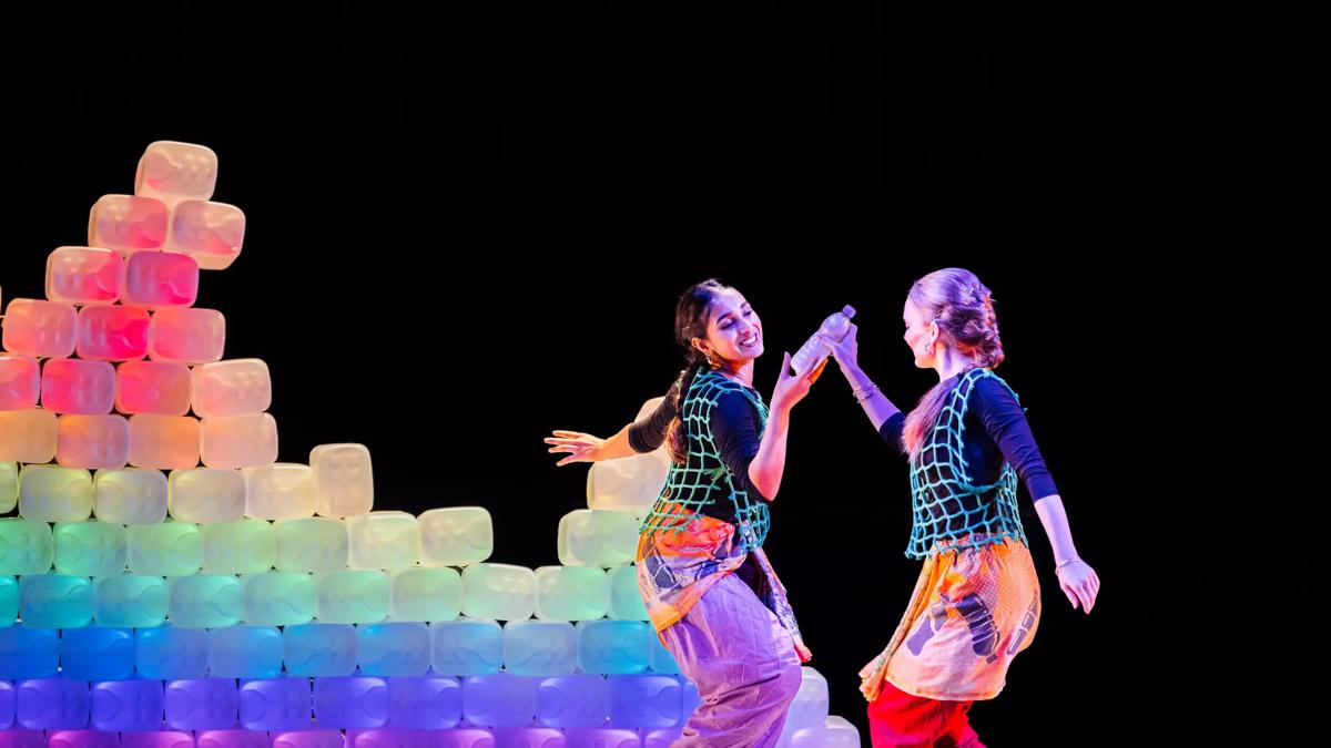 Two female performers dance together in front of a rainbow light wall