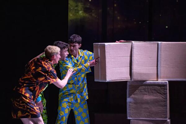 Three actors wearing colourful patterned clothes looking at a stack of boxes.