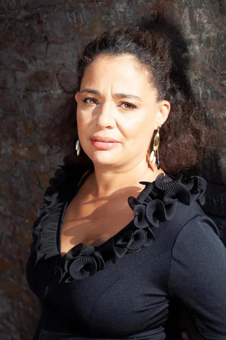 Half-length portrait of Karen McCarthy Woolf, who has a serious face and wears a black ruffled top and pendant earrings