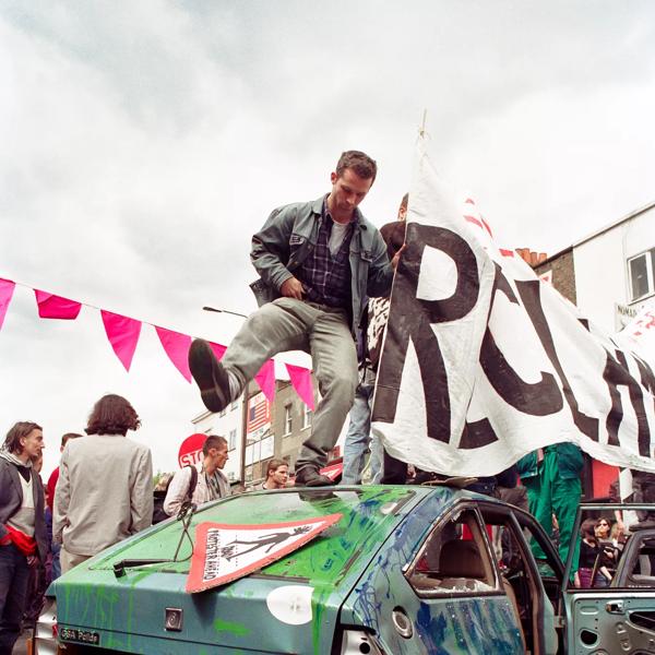An activist stands on the bonnet of a green truck holding a white protest sign that says 'Reclaim'. He wears a leather jacket and jeans and appears to be stomping on top of the car. 