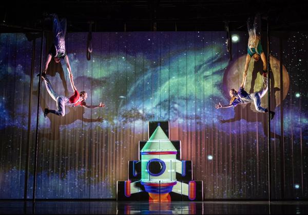 Actors suspended in the air with a galaxy in the background and a spaceship below them.
