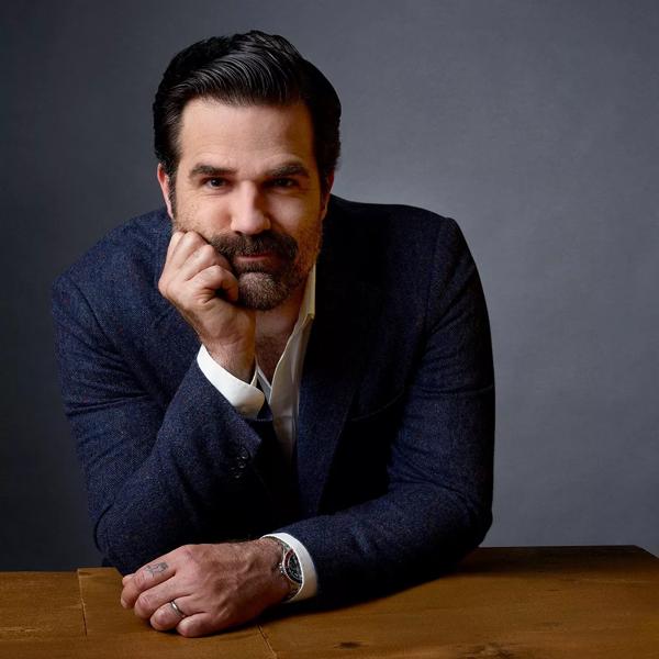 Portrait of comedian and author Rob Delaney. He leans on a wooden table in front of a grey background. He wears a blue suit jacket over a white shirt and leans his chin on his right hand.  
