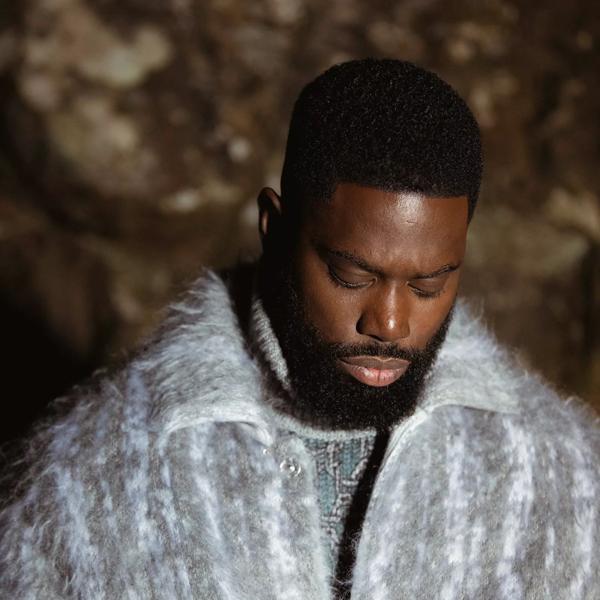 Ghetts stands against a rocky background wearing a light blue jacket.