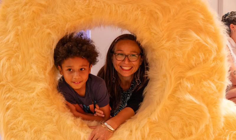 A woman and child poking their heads through a furry orange doughnut-shaped circle