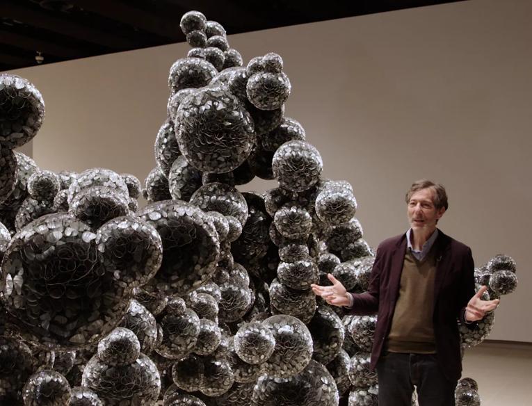 Ralph Rugoff, an older White man, wearing a suit, stands next to the sculptural work 'Untitled (Mylar)' (2011), which consists of spheres made of thousands of folded discs of mylar, part of When Forms Come Alive at Hayward Gallery.