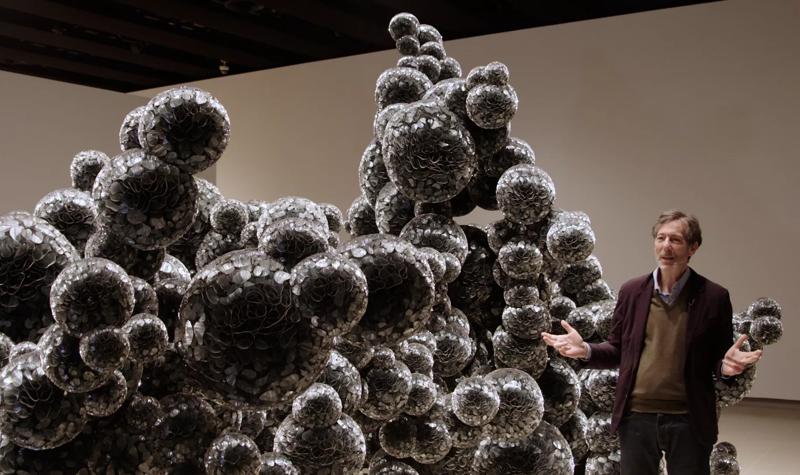 Ralph Rugoff, an older White man, wearing a suit, stands next to the sculptural work 'Untitled (Mylar)' (2011), which consists of spheres made of thousands of folded discs of mylar, part of When Forms Come Alive at Hayward Gallery.