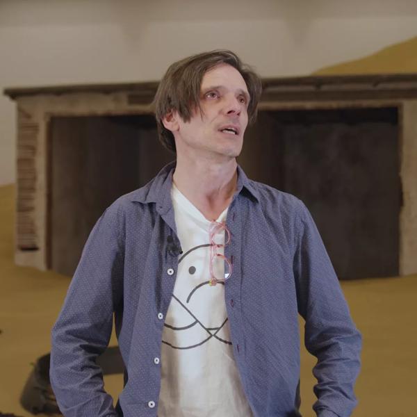 The artist Jeremy Deller wearing an open blue shirt over a white t-shirt stands inside the Hayward Gallery in front of a work by Mike Nelson, part of the Mike Nelson: Extinction Beckons exhibition at Hayward Gallery