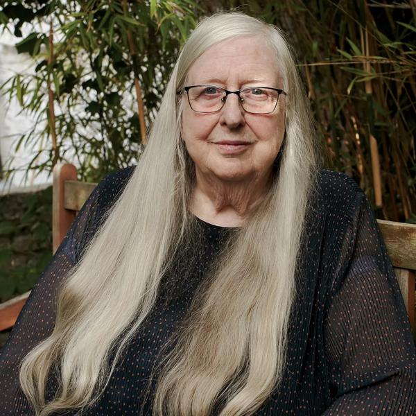The poet Eilean Ni Chuilleanain, an older White woman with very long fair hair, is seated outside and wears a black top and glasses