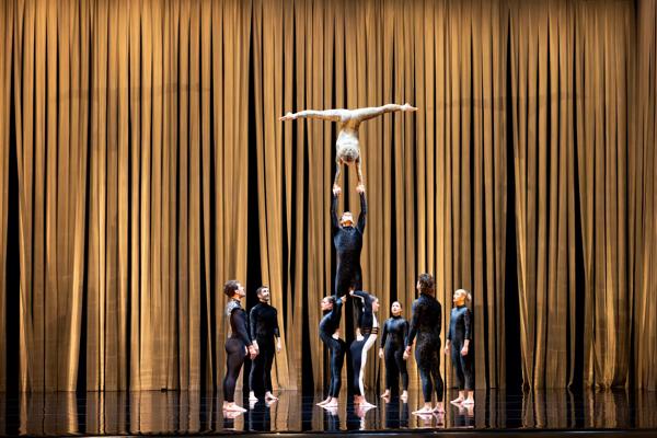 Two performers are balancing another performer on their backs who is balancing a person dressed in a gold leotard upside down