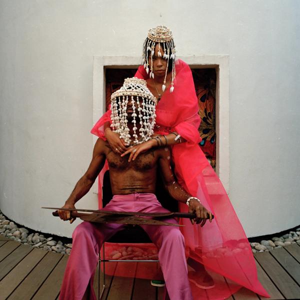 A topless Black man sits, behind him a Black woman stands with her arms around his shoulders. Both wear red and hats which trail over their faces