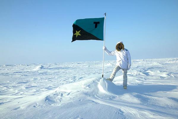 Artist Tavares Strachan, a Black man in his 30s, stands at the North Pole on a white landscape of ice and snow, wearing an all white snowsuit. He is holding a flag pole, on top of which is a flag that is aquamarine and black divided diagonally with a Gold star on the lower black half and a black 't' on the upper aquamarine half