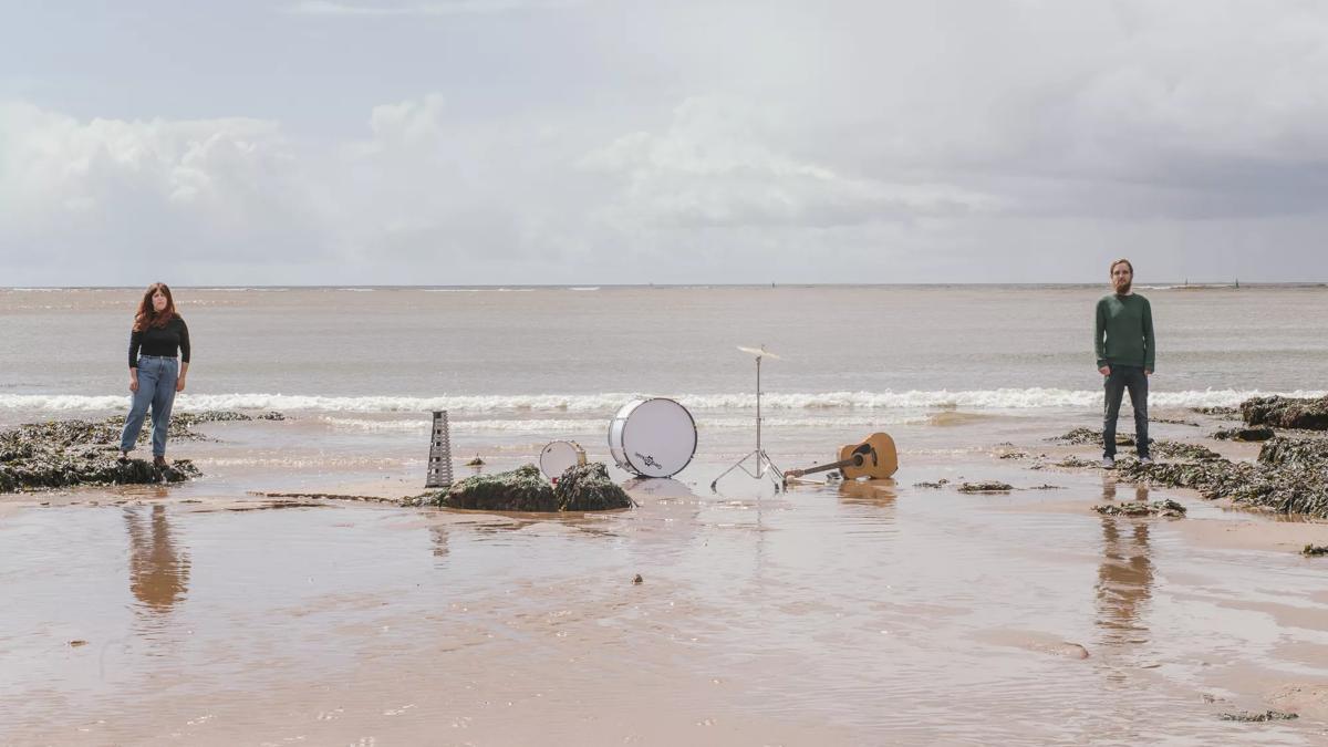 Jack Dean & Company (one man and woman) stand on opposite sides of a beach scene. Inbetween them instruments are scattered on the sand. A guitar, a kick drum, a snare and a high hat. In front of the drums obscuring them slightly is a pile of green seaweed.