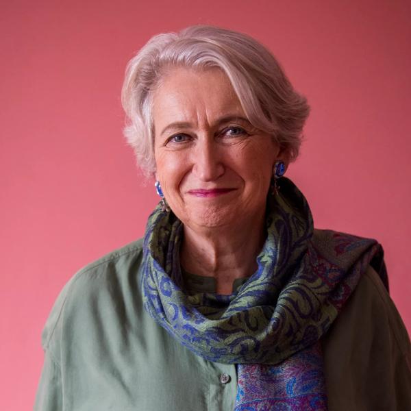 Lindsey Hilsum stands against a pink background wearing a green jumper and scarf.