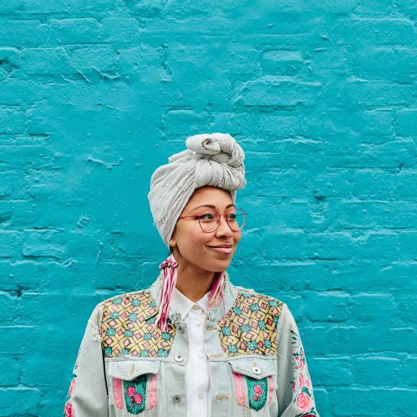 Yassmin Abdel-Magied stands against a turquoise wall wearing pink earrings, a grey headscarf and a colourful jacket