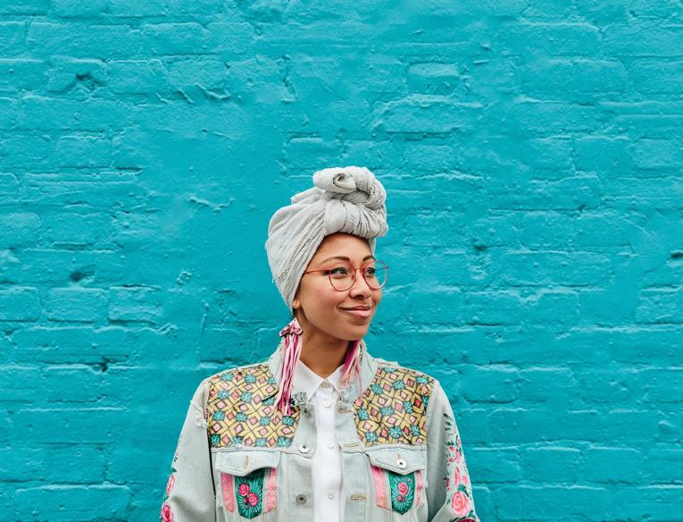 Yassmin Abdel-Magied stands against a turquoise wall wearing pink earrings, a grey headscarf and a colourful jacket