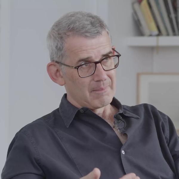 Edmund de Waal, a white man with grey hair, wearing glasses and a dark shirt, talks to camera whilst seated in his studio, behind him shelves are visible, one of which contains books