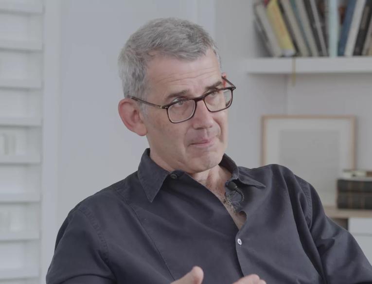 Edmund de Waal, a white man with grey hair, wearing glasses and a dark shirt, talks to camera whilst seated in his studio, behind him shelves are visible, one of which contains books