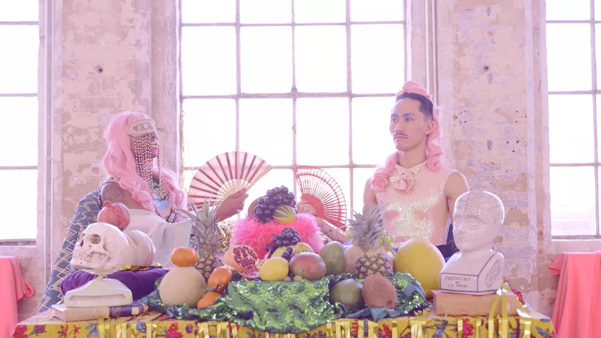 Theatrical setting with two people waring a pink wig and a holding a fan. They are behind a table with section of fruits and sculpture heads.