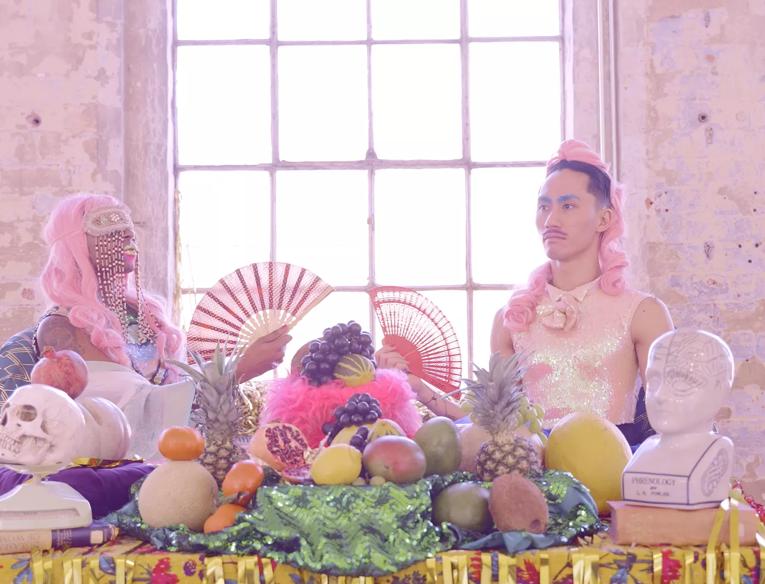 Theatrical setting with two people waring a pink wig and a holding a fan. They are behind a table with section of fruits and sculpture heads.