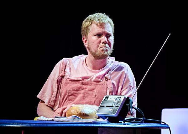 Person wearing pink dungarees and a pink T-shirt sits at a table with a loaf of bread and a radio on top of it.