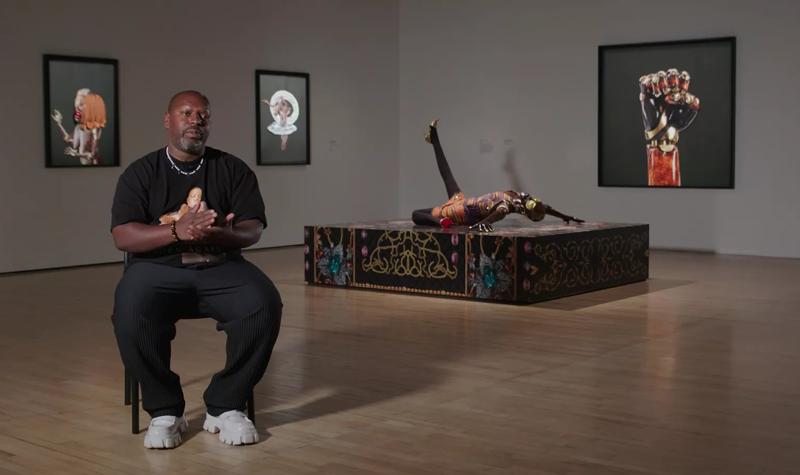 Artist Rashaad Newsome seated inside Hayward Gallery, in the back ground are some of his works within In the Black Fantastic including threee wall-mounted collage pieces and a sculpture